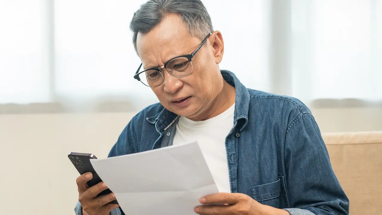 Mature Asian old man is stressed because he looks at many expense bills and no money to pay them holding cell phone. Not enough money on credit card to pay. Senior old man with bill and smartphone stock photo