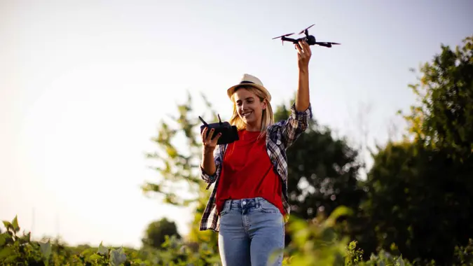 New generation of young woman with organic vegetable business, blonde girl on a raspberry field wearing casual clothing and hat, exploring the integrity of organic vegetables using drone.