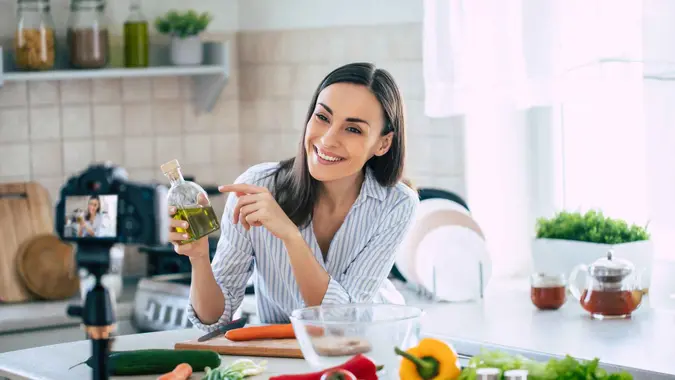 Professional beautiful happy young woman is blogging for her kitchen channel about healthy living in the kitchen of her home and looking on camera on a tripod.