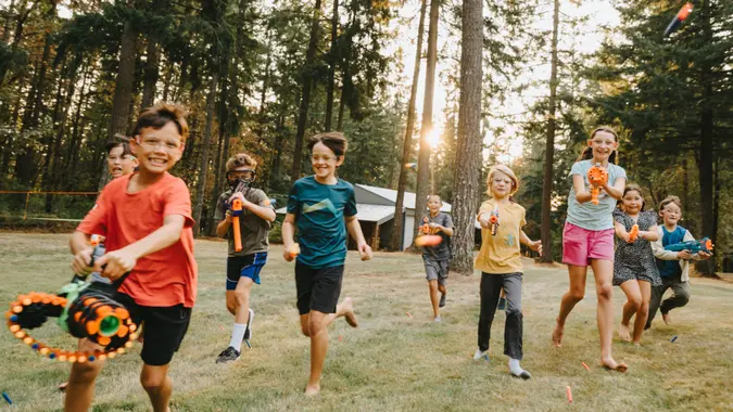 A multiracial group of elementary age and tween boys and girls run and shoot nerf guns toward the camera while having a fun nerf gun battle in a grassy field surrounded by fir trees.