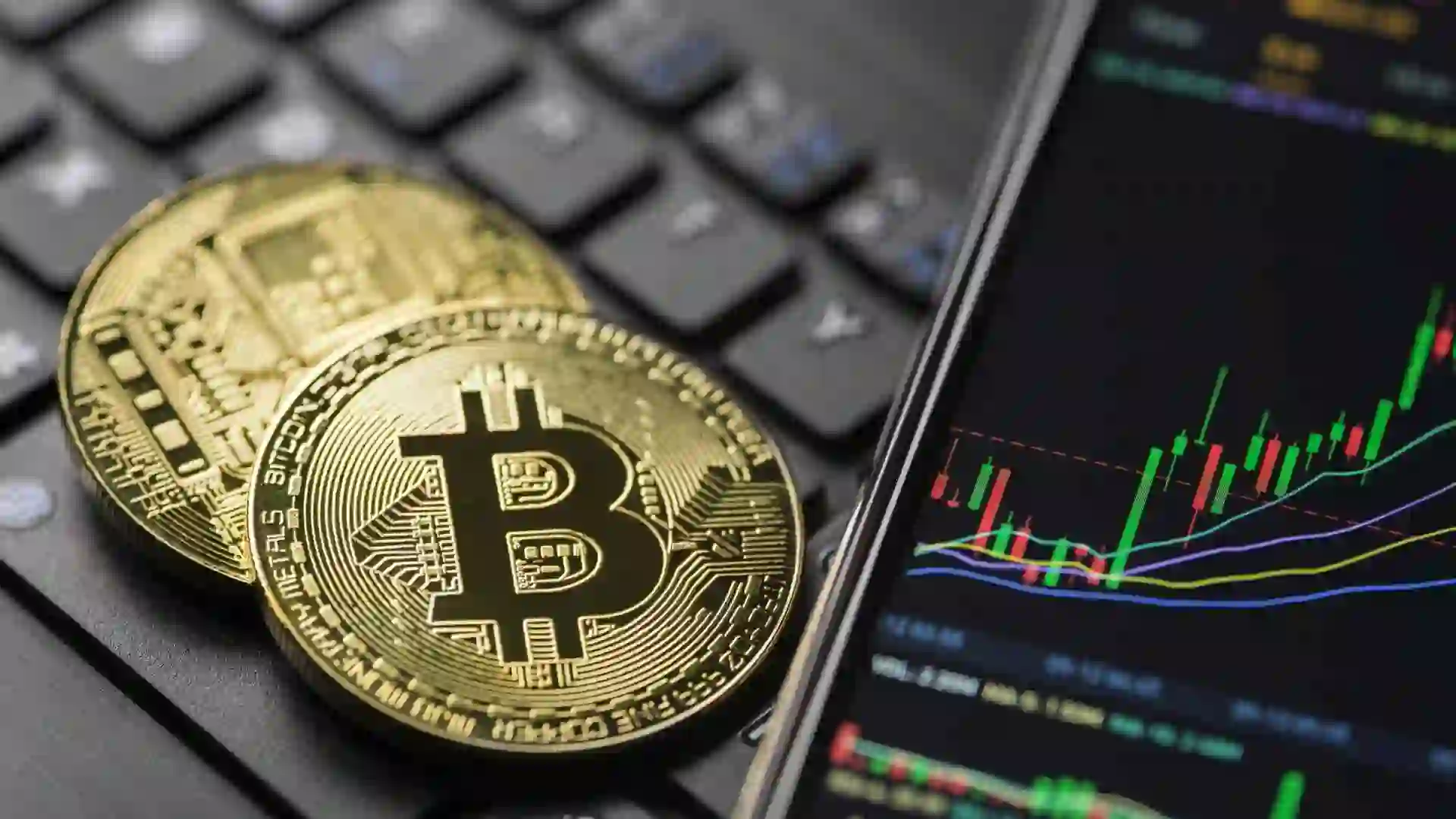 Two gold coins with the bitcoin symbol on a computer keyboard next to a smartphone displaying a stock chart.
