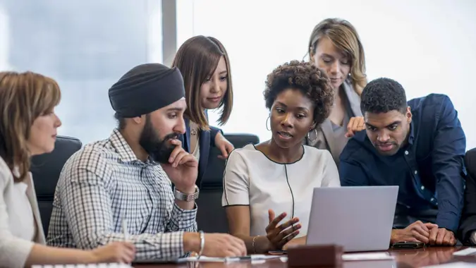 A diverse group of business people gather around a laptop in a modern office and discuss what they see.
