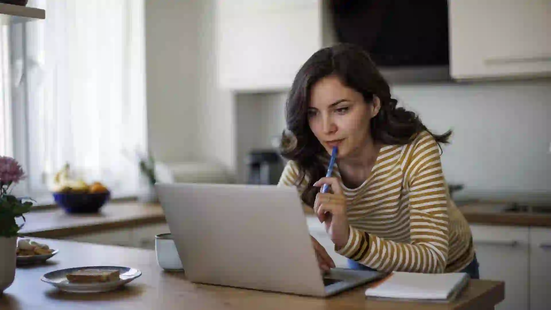 A focused young woman with long, wavy hair works on her laptop, which is propped up on her modern kitchen counter.