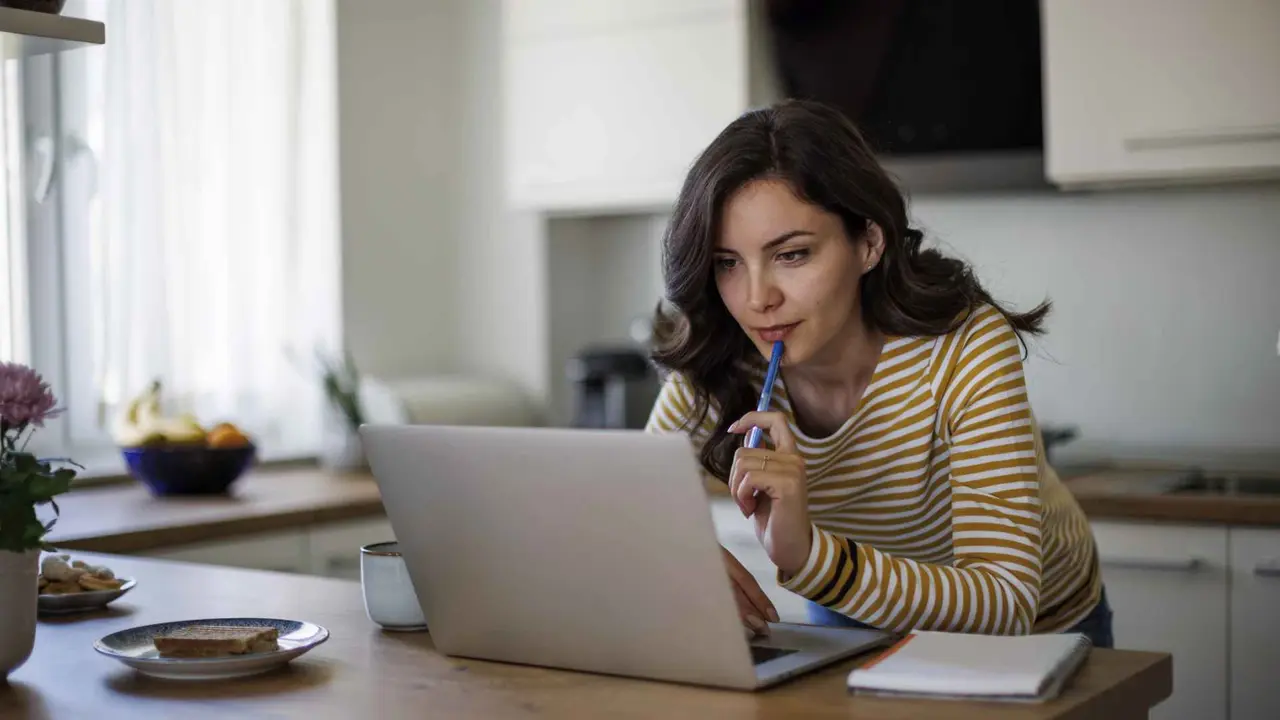 A focused young woman with long, wavy hair works on her laptop, which is propped up on her modern kitchen counter.