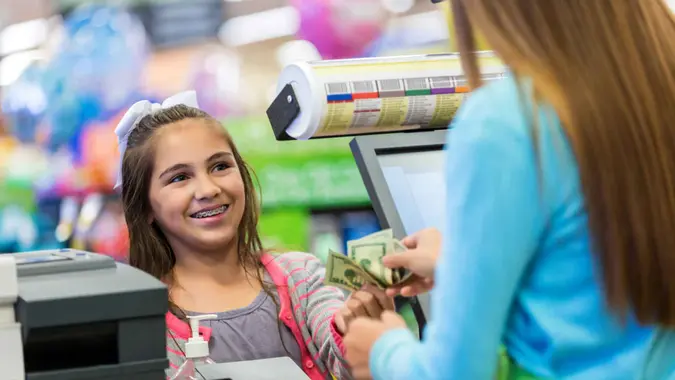 Elementary age Caucasian little girl with brown hair and braces is smiling while paying for a purchase at checkout counter in local store or supermarket.