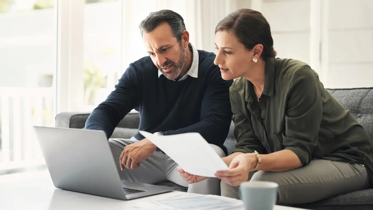 Couple sitting in their living room in front of a coffee table, reviewing their finaces while looking at a computer.
