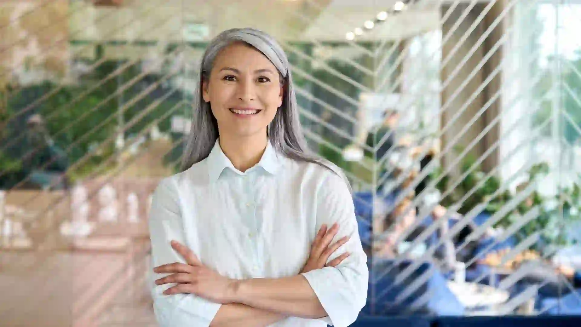 Happy smiling confident middle aged Asian older senior female leader businesswoman standing in modern office workplace looking at camera arms crossed.