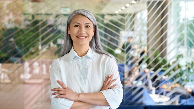 Happy smiling confident middle aged Asian older senior female leader businesswoman standing in modern office workplace looking at camera arms crossed.
