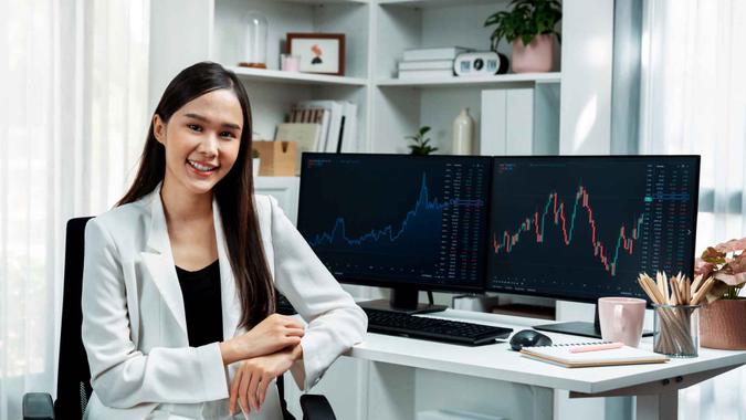 Profile of young Asian businesswoman smiling on happy face with white suit sitting chair against dynamic stock exchange market investing graph on pc showing screen on desk at modern office.