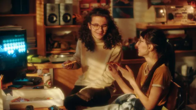 Teens eating pizza and talking to each other after playing game during their leisure time in furnished garage.
