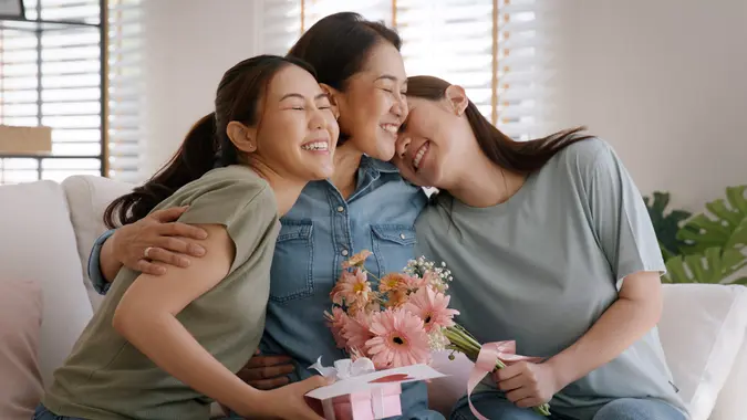 Mother day two grown up child cuddle hug give flower gift box red heart card to mature mum. Love kiss care mom asia middle age adult three people sitting at home sofa happy smile enjoy family time. stock photo