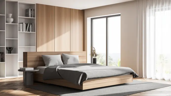 Corner of stylish master bedroom with white and wooden walls, wooden floor, comfortable king size bed, a bookcase and windows with blurry view.