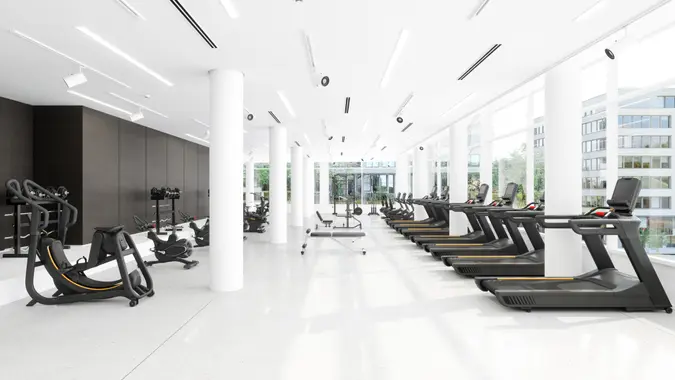 Modern Gym Interior With Treadmills, Exercise Bikes, Sports Equipments And City View Through Window.