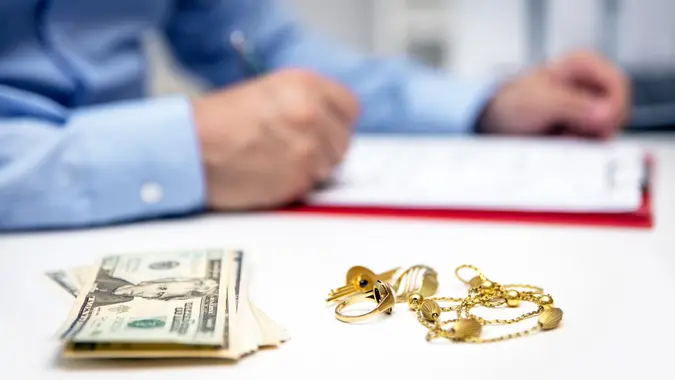 Close-up of gold jewelry and cash in foreground with businessman in background, representing selling valuables.