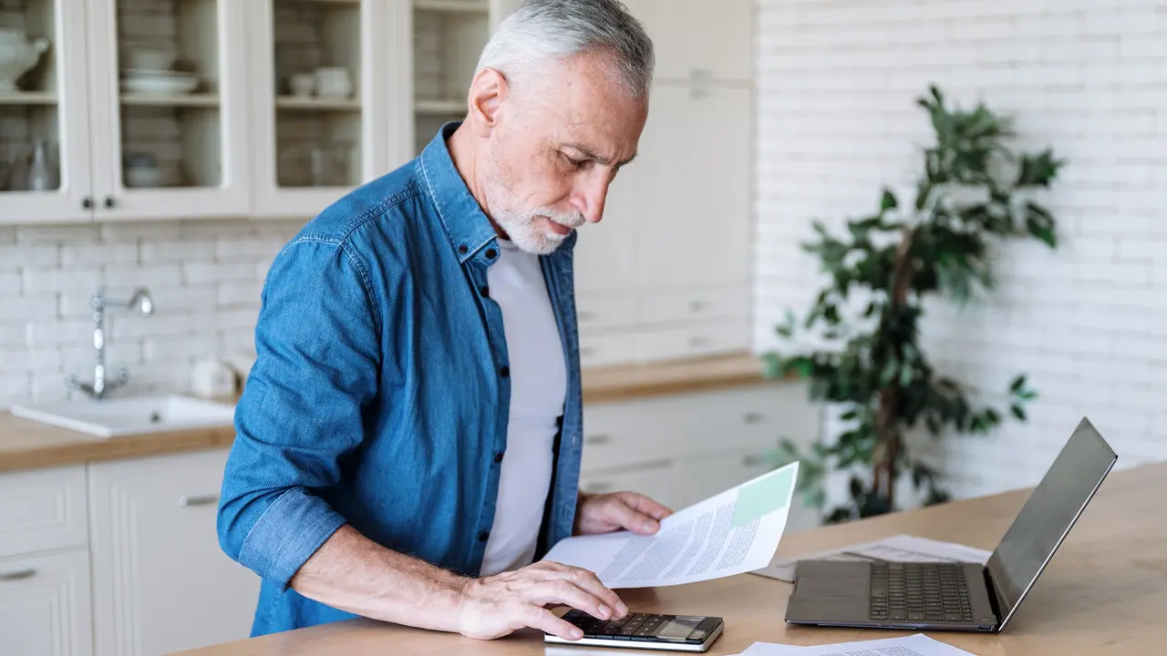 Focused senior man using a calculator and a laptop.