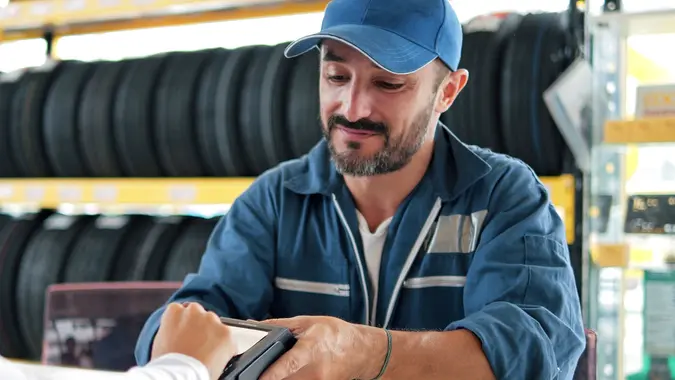 Car mechanic man in blue uniform hold payment terminal for paying with credit card while owner driver customer using credit card touching for pay at vehicle repair shop stock photo