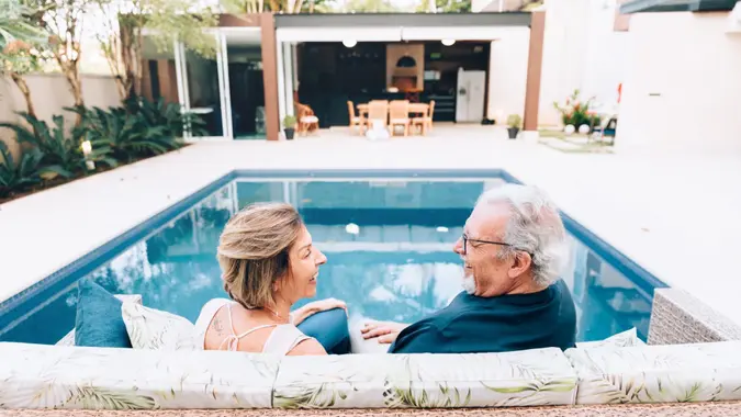 A happy senior couple sits on a couch outside, in front of a pool.