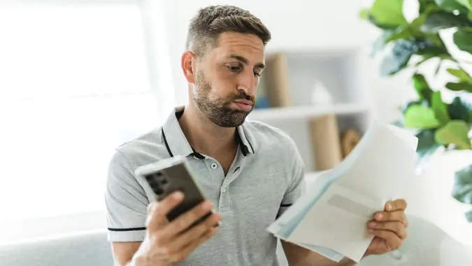 A middle-aged man with a furrowed brow and anxious expression reads from a surprising paper document in his hands.