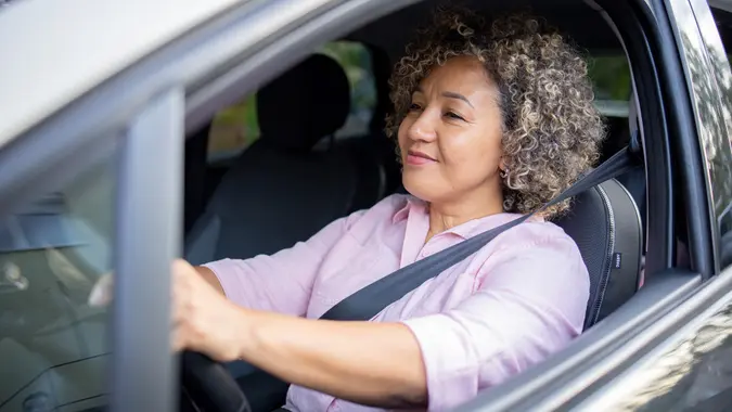A smiling woman with curly hair in a pink button-down shirt is driving a car on her way to work