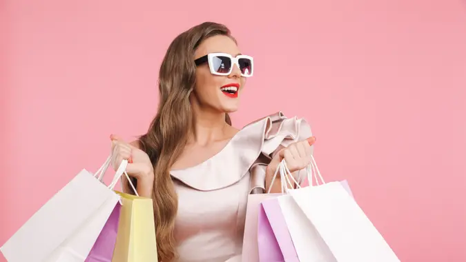 Satisfied charming woman with long brown hair wearing dress and stylish sunglasses holding colorful shopping bags with smile isolated over pink background.