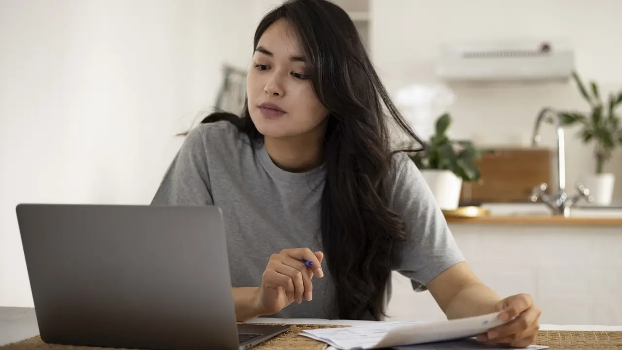 A thoughtful woman managing her personal finances at home, surrounded by documents and a laptop.