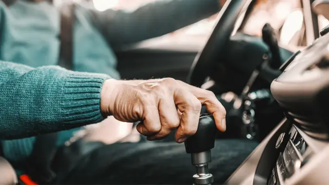 An older person in a car. Close-up of a hand on a gear shift.