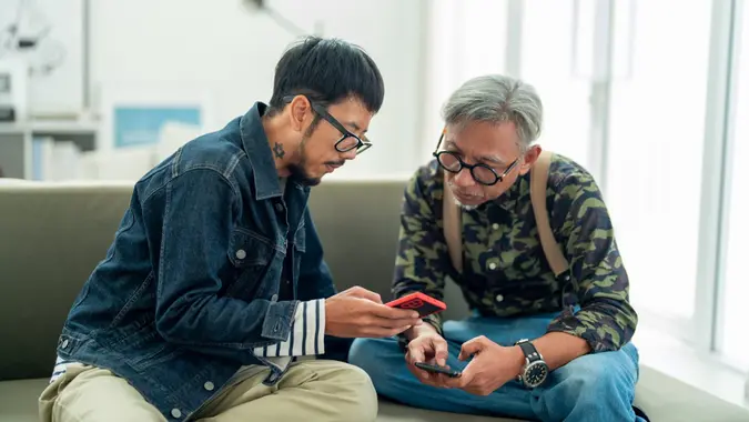 Asian Man and Senior Father Enjoying Quality Family Time Using Smartphones in Cozy Living Room.