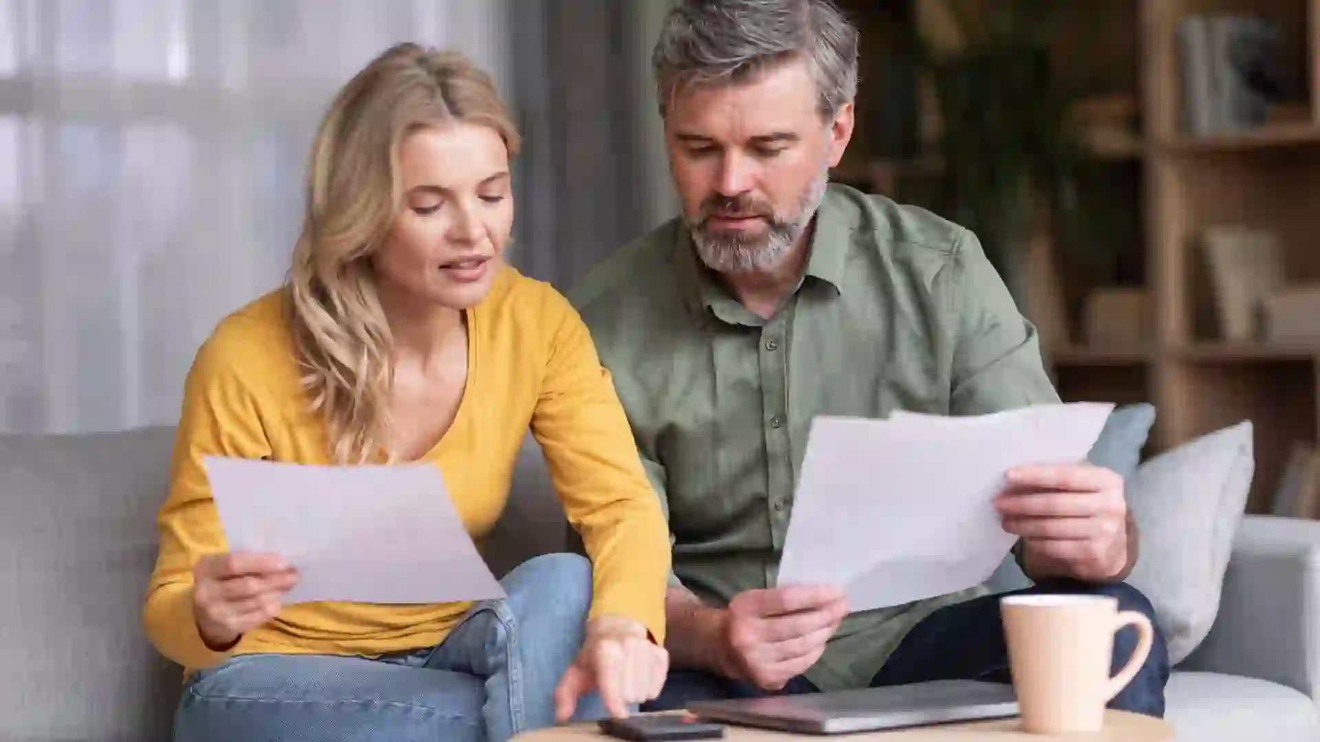 Middle aged husband and wife sitting on a couch holding papers and calculating family budget together