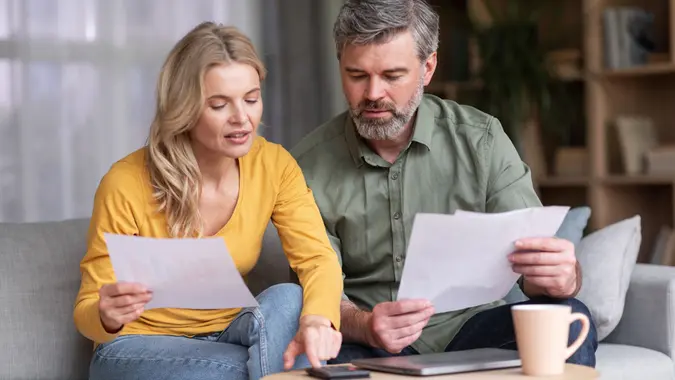 Middle aged husband and wife sitting on a couch holding papers and calculating family budget together