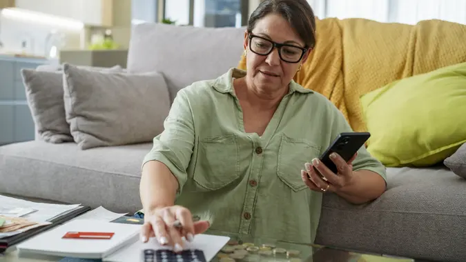 Woman sitting on couch using calculator and mobile phone checking expenses and bills and planning home finances stock photo