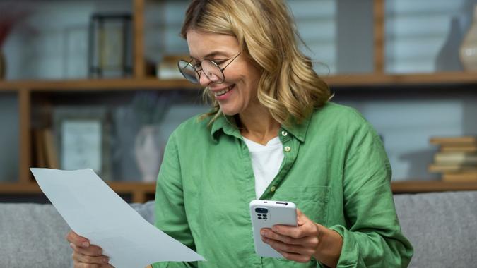 An elderly smilling woman sits at home on the sofa, holds the phone in her hands, works with documents, financial accounts, rent, loans, agreements stock photo