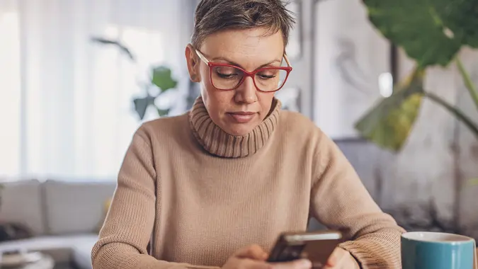 Shot of a woman with glasses on sitting down while using her smartphone to review her financials.
