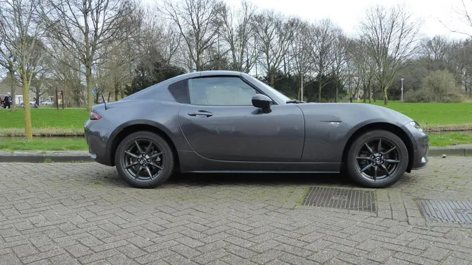 A dark gray Mazda MX-5 Miata sitting in a driveway with trees in the background.