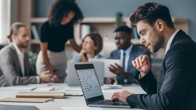 Confident businessman sitting at the office desk and using laptop while colleagues working on background stock photo