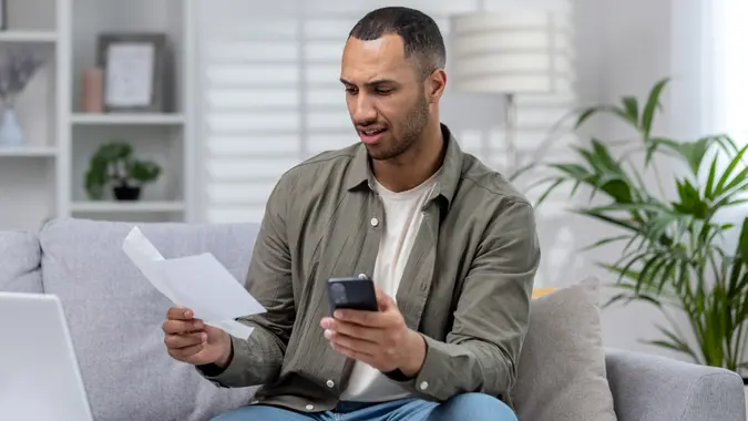 Young hispanic businessman working remotely at home on laptop. Holding the phone and anxiously looking at documents, checking accounts, financial problems and bankruptcy stock photo