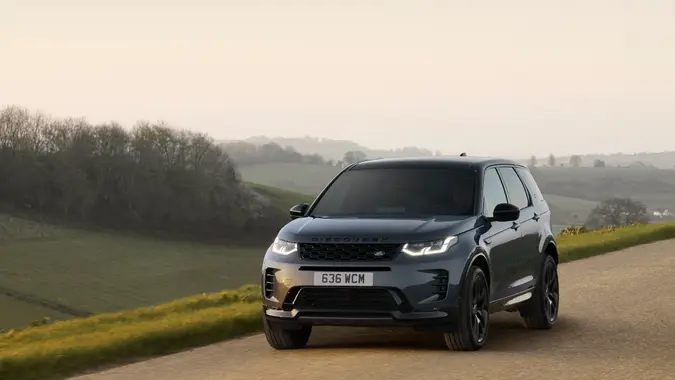A black 2024 Land Rover Discover Sport driving along a road with trees in the background.
