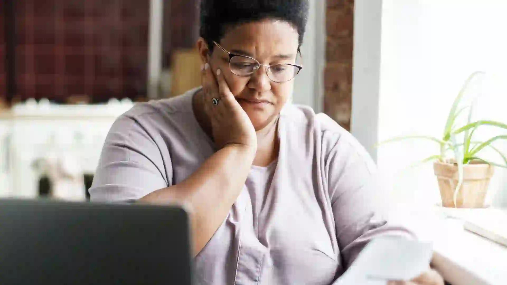 A mature African American woman in glasses touches her cheek, looking sad and puzzled by a high utility bill.
