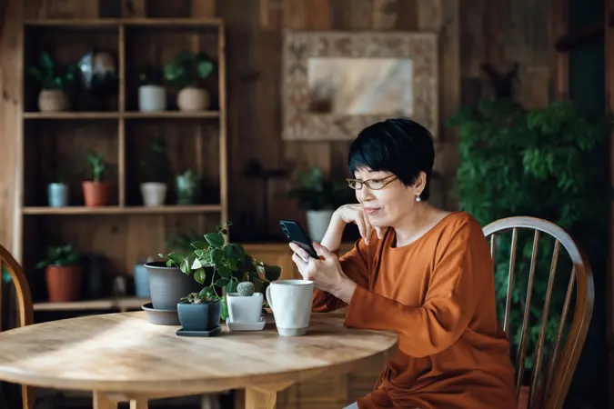 Smiling senior Asian woman sitting at her table in the dining room while using a smartphone in her left hand.