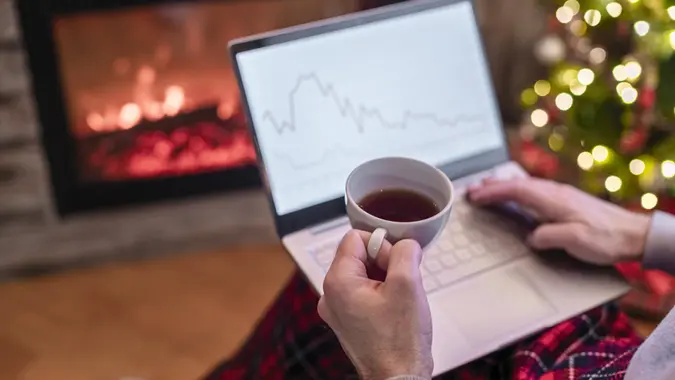 Man freelancer working on laptop with graphs and charts on screen sitting near christmas tree and fireplace with cup of tea.