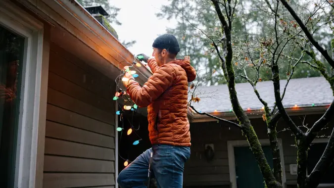 A middle aged Caucasian man works on his Christmas holiday decor, either putting up or taking down the colored light bulb decorations from off of his house gutters.