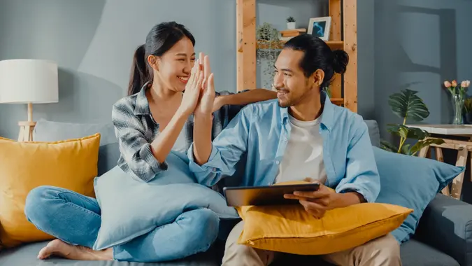 A happy couple is sitting on a couch at their home, high-fiving each other and using a tablet.