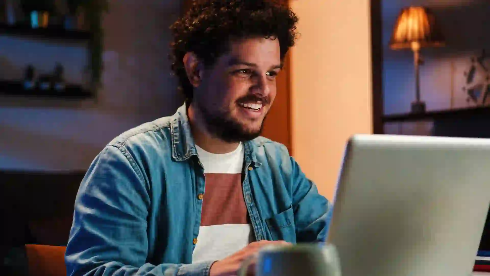 A Young, happy Man working at his home with a laptop open in front of him on a desk at late night.