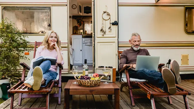 Portrait of senior married caucasian couple having rest on the deck chairs with food and wine just against their caravan home.
