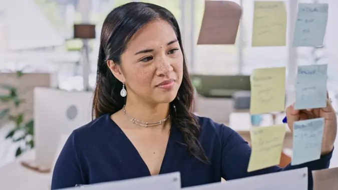 Woman, thinking or glass board with sticky notes for brainstorming, planning and project management in modern office. Young person, thought or scrum with idea on wall, creative strategy or workplace stock photo