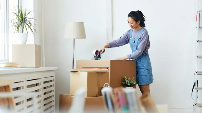 Side view portrait of young Asian woman packing cardboard boxes and smiling happily while moving to new home or apartment, copy space.