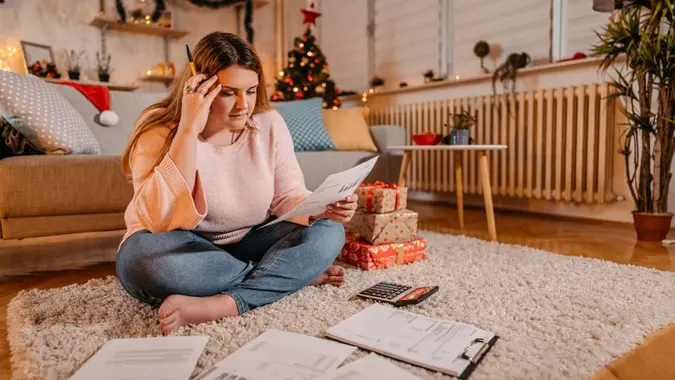 A young woman with a worried expression sits at a table, calculating her bills amidst festive Christmas decor.