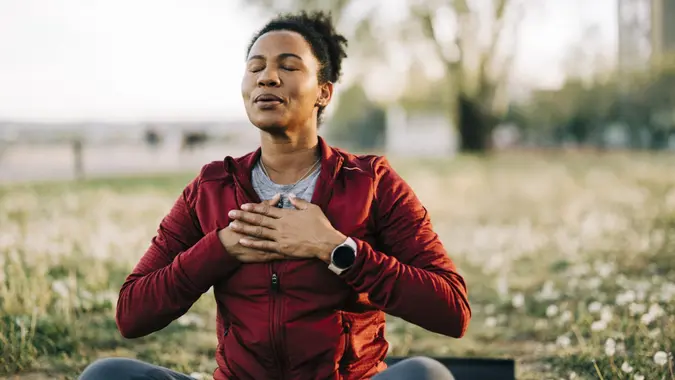A woman sitting in a field with her eyes closed and her hands on her chest, meditating.