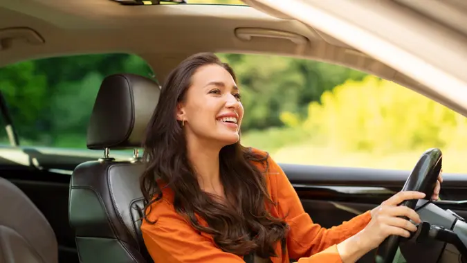 Joyful European woman driving brand new car, looking at rear view mirror and smiling, enjoying her new auto, side view, copy space.