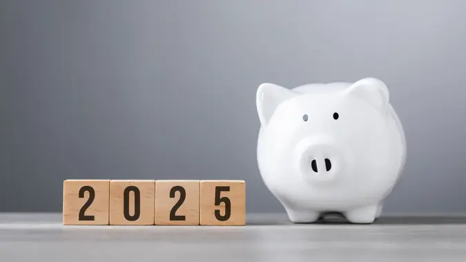 2025 written on a wooden block next to a white piggy bank placed on a wooden table against a bright white.