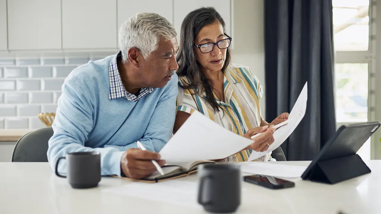 An older couple sitting in their brightly lit kitchen at home, reviewing their finances on paper together